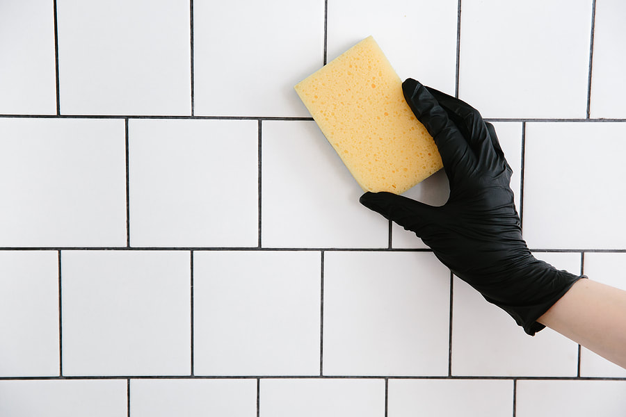 a hand in black gloves holding a sponge on a white tiled wall
